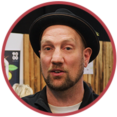 Ein Mann mit leichtem Bart und Schnurrbart trägt einen schwarzen Fedora-Hut und eine schwarze Jacke und steht in einem Innenbereich vor Holzpaneelen und Plakaten.