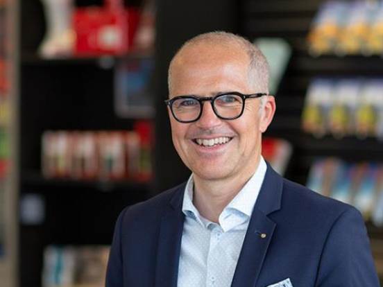Ein lächelnder Mann mit Brille und marineblauem Blazer steht in einem Innenraum, mit bunten, verschwommenen Regalen im Hintergrund.