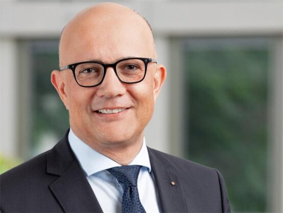 Ein lächelnder Mann mit Glatze, schwarzgerahmter Brille, dunklem Anzug, weißem Hemd und blauer Krawatte steht vor einem unscharfen Innenhintergrund.